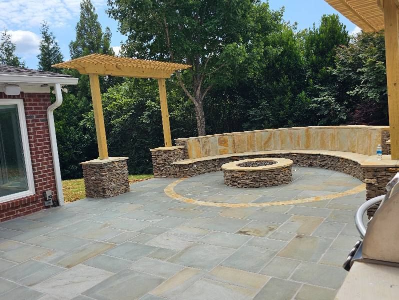 Patio with fire pit, stone seating, and pergola under a blue sky.