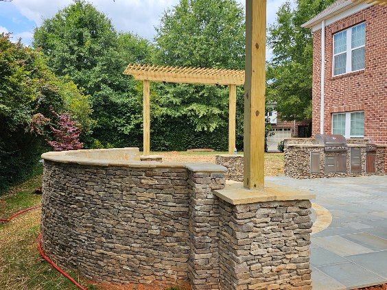 Stone patio with a wooden pergola, brick wall of a house visible.