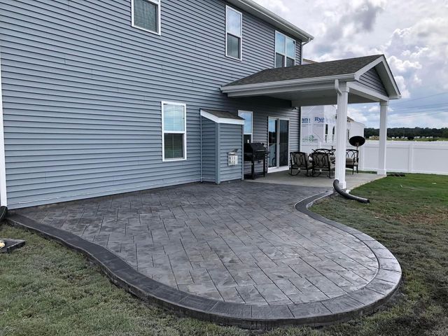 Gray stamped concrete patio with dark border, attached to a gray house, next to a grassy yard.