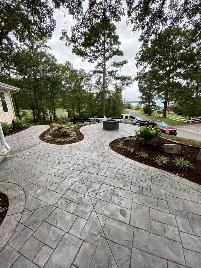Stone patio with curved edges, landscaped beds with plantings. Trees and a water view in background.