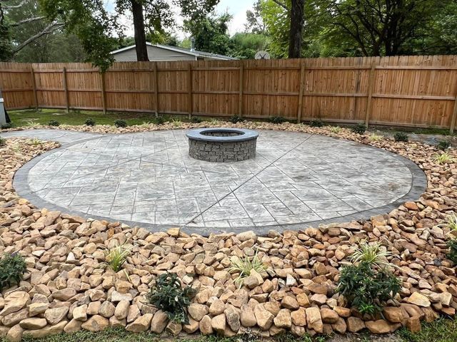 Circular stone patio with fire pit, bordered by rocks and landscaping, fenced backyard.