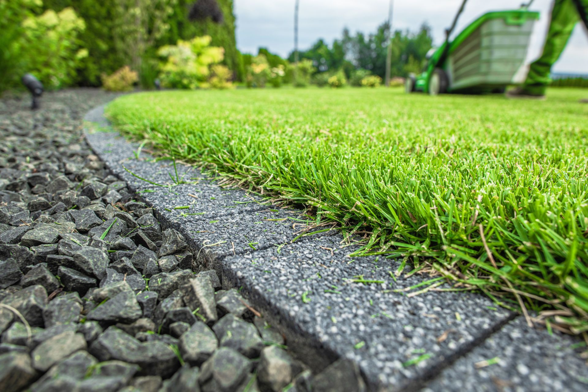 Lawn edge with gray pavers and gravel, bright green grass, and a lawnmower in the background.