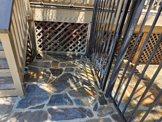 Stone steps leading to a latticework area, a black metal gate to the right, beneath a wooden deck.