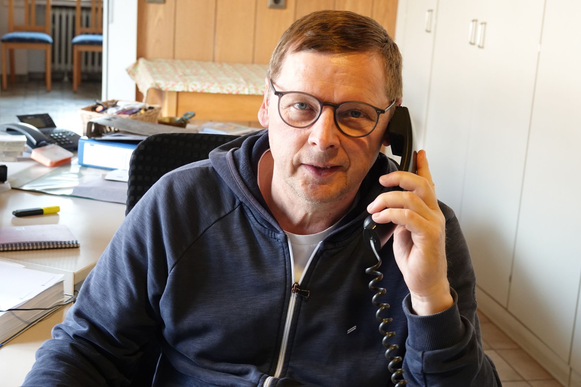 Ein Mann sitzt an einem Schreibtisch und hält ein Telefon in der Hand. Im Hintergrund sind Papiere und Schränke zu sehen.