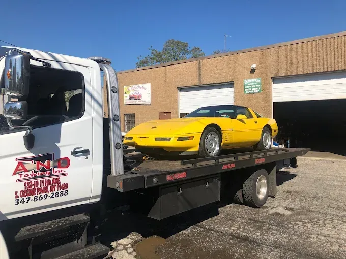 Yellow Corvette on a flatbed tow truck in front of a brick building on a sunny day.