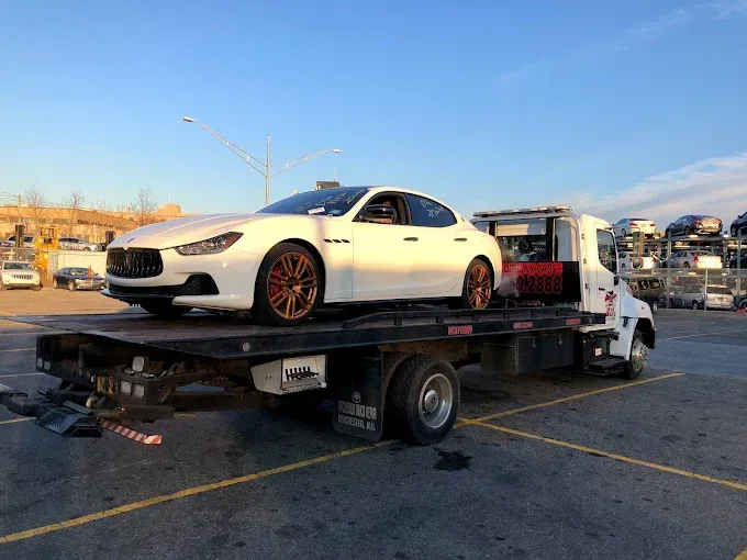 White Maserati sedan on a flatbed tow truck in a parking lot.