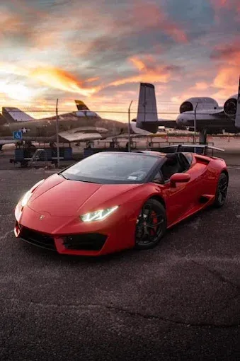 Red Lamborghini convertible parked in front of vintage airplanes at sunset.