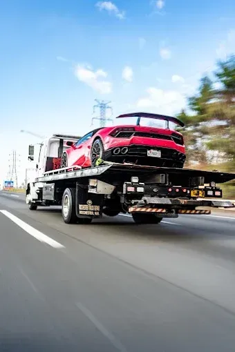 Red Lamborghini sports car on a flatbed tow truck traveling on a highway under a blue sky.