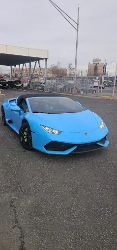 Blue Lamborghini parked on wet pavement near an industrial lot at dusk