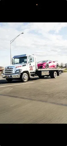 Tow truck carrying a pink sports car on a paved road under a partly cloudy sky.