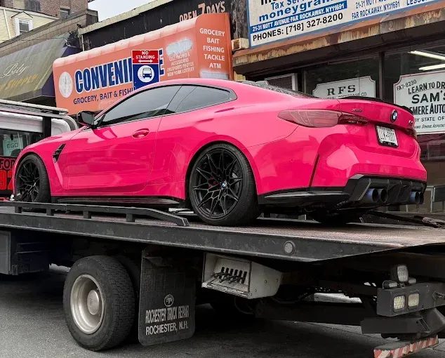 A bright pink BMW sports car on a tow truck, with a city backdrop.