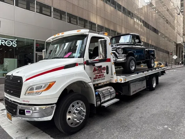 White tow truck carrying a black vintage truck on a city street.