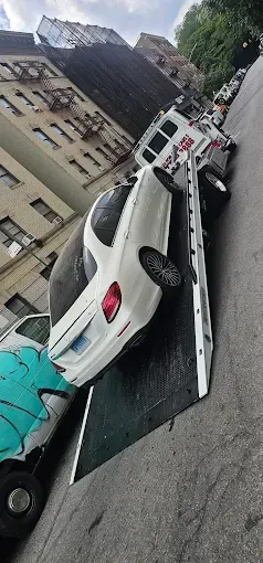 White car on a tow truck on a steep city street. Buildings line the street.