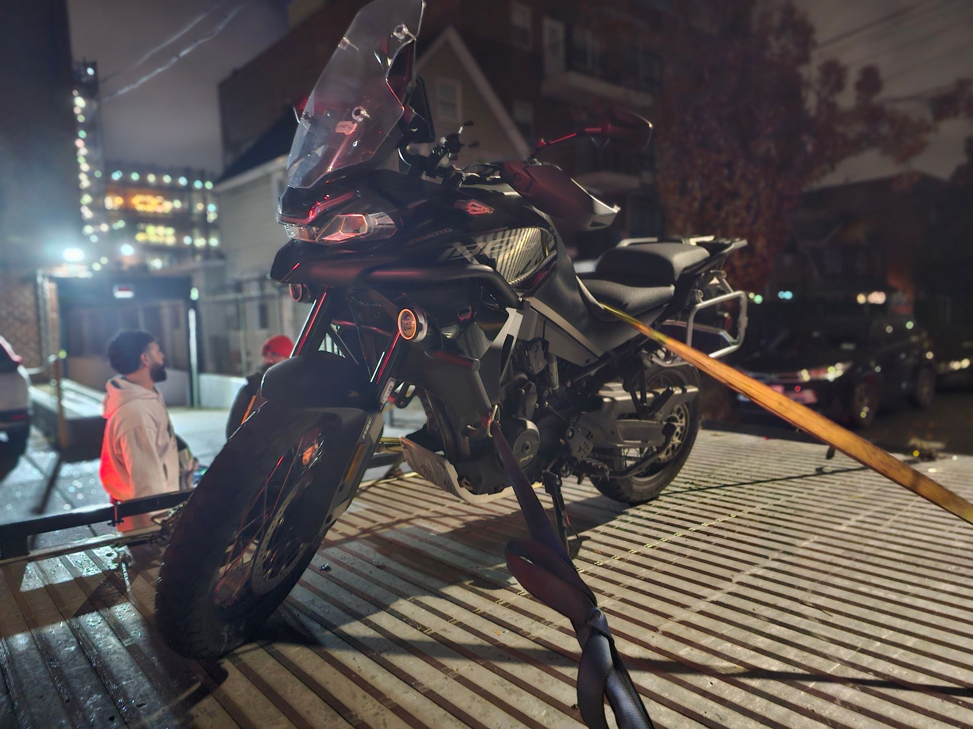 Motorcycle on a tow truck at night. Black motorcycle strapped down. Man watches in the background.