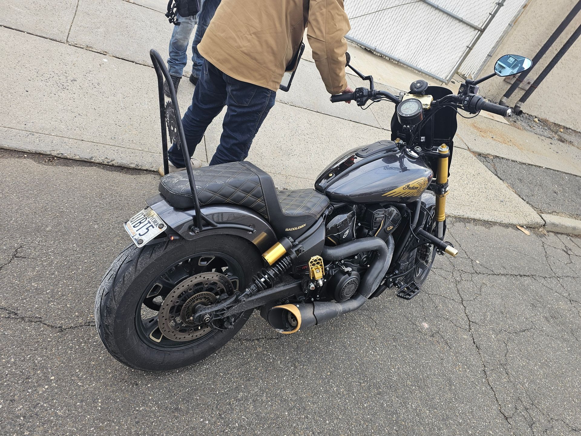 Black custom motorcycle with gold accents parked on pavement. A person in jeans and a brown jacket stands nearby.