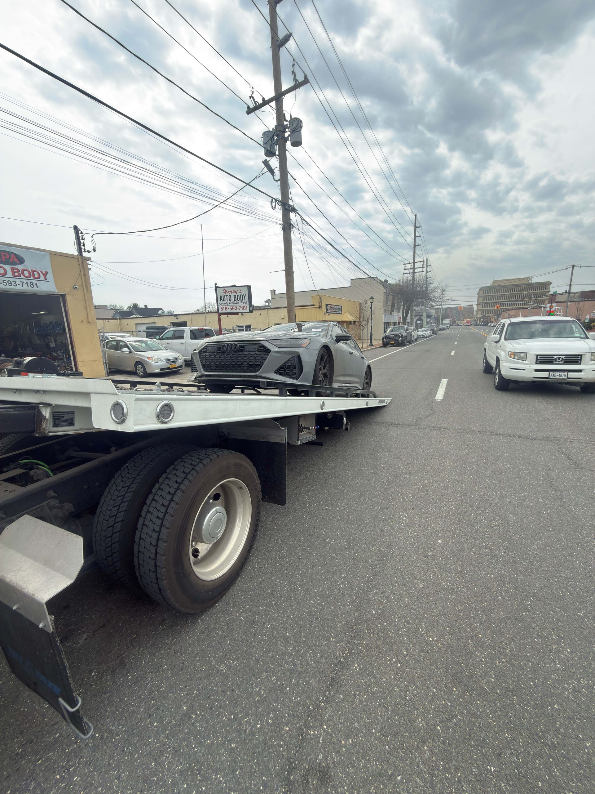 Tow truck carrying a car on a roadside with utility poles and traffic under cloudy skies