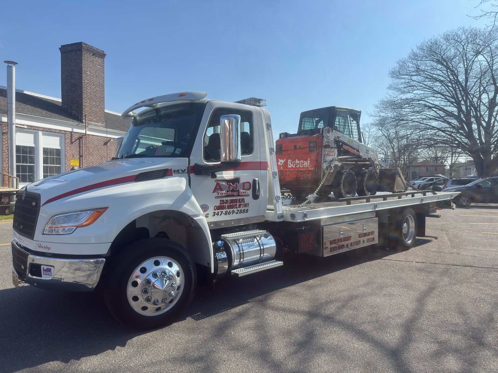 Tow truck carrying a red pickup on a flatbed, parked outside a brick building on a sunny day