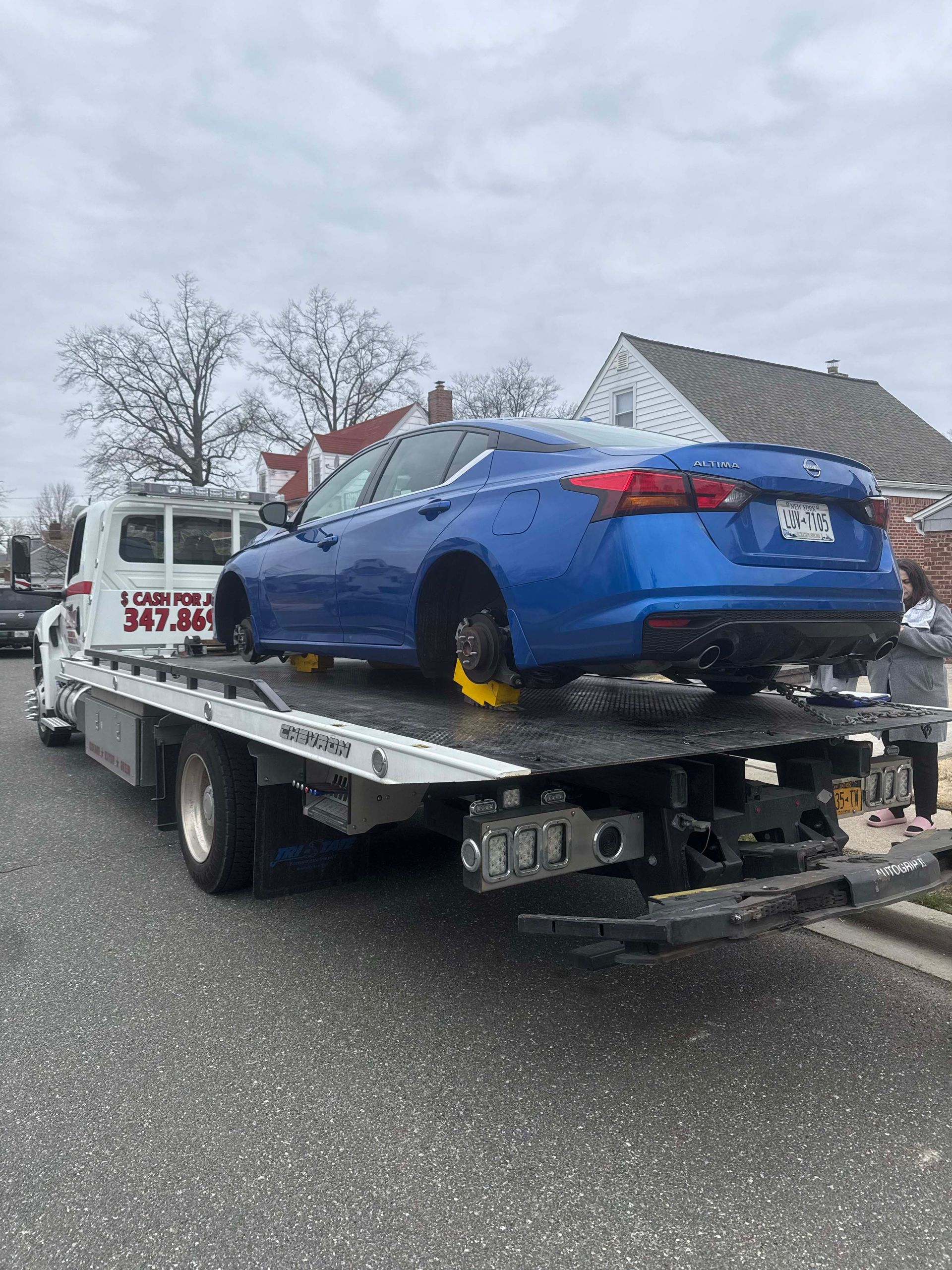 Blue sedan on a flatbed tow truck with yellow wheel locks, parked outdoors on a cloudy day