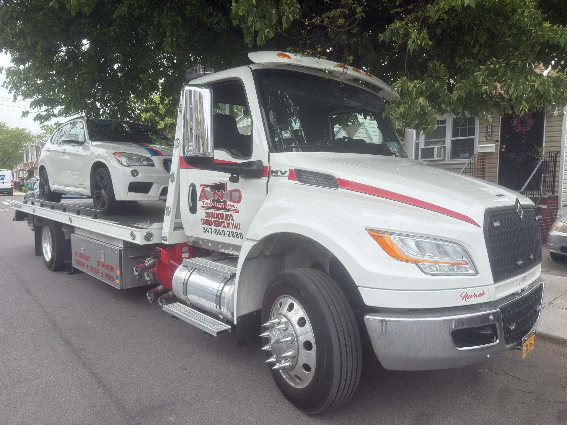 White tow truck carrying a white car on a flatbed under trees by the curb
