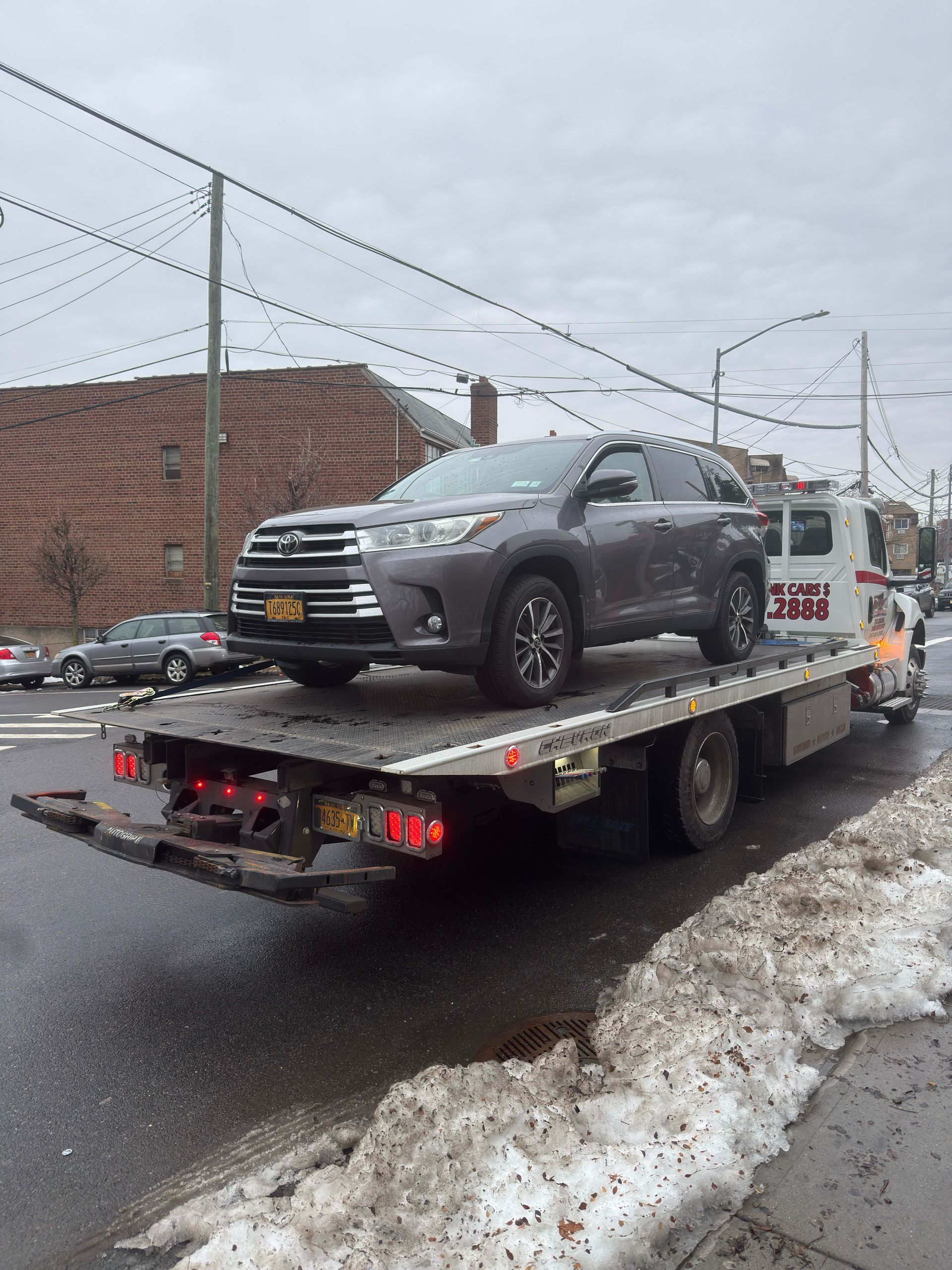 Gray SUV on a tow truck flatbed in a snowy lot beside a brick building and chain-link fence