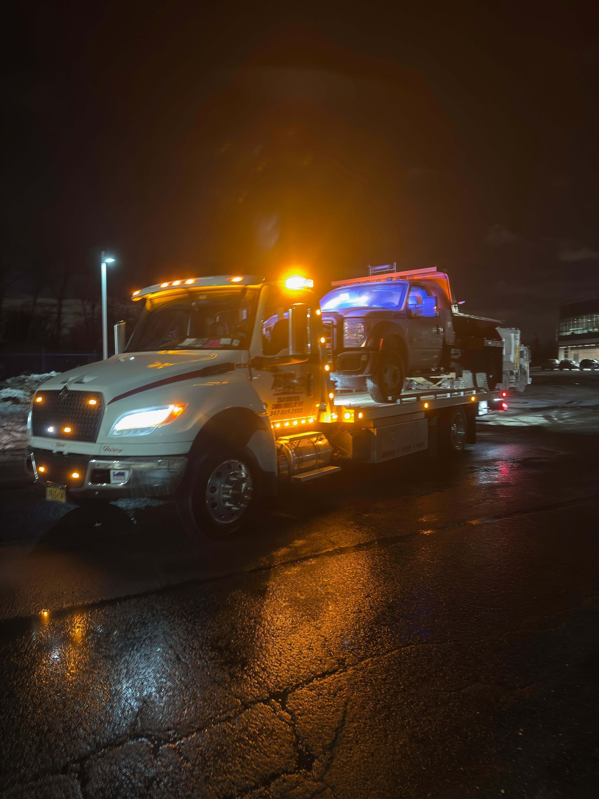 Tow truck with flashing amber lights on a wet street at night, carrying a car on its flatbed