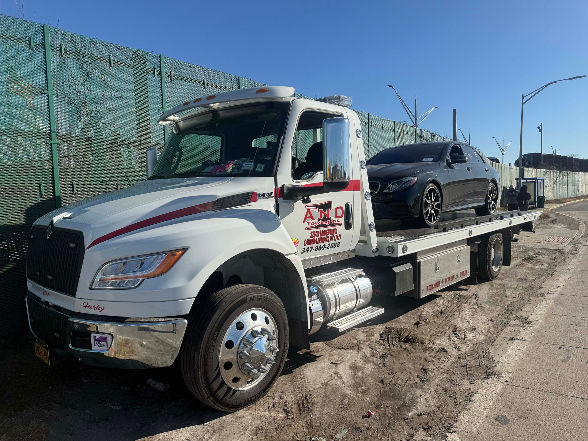 Tow truck carrying a black car on a flatbed beside a roadside fence under clear sky