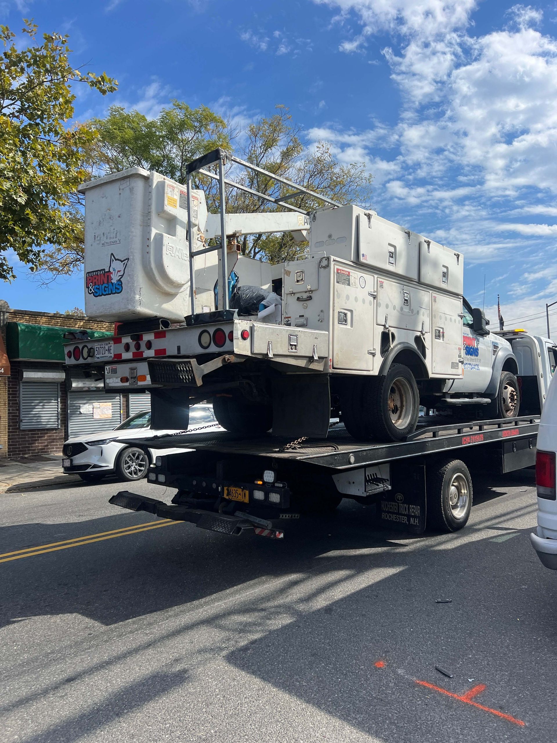 Tow truck hauling a large white utility truck on a flatbed in a sunny parking lot