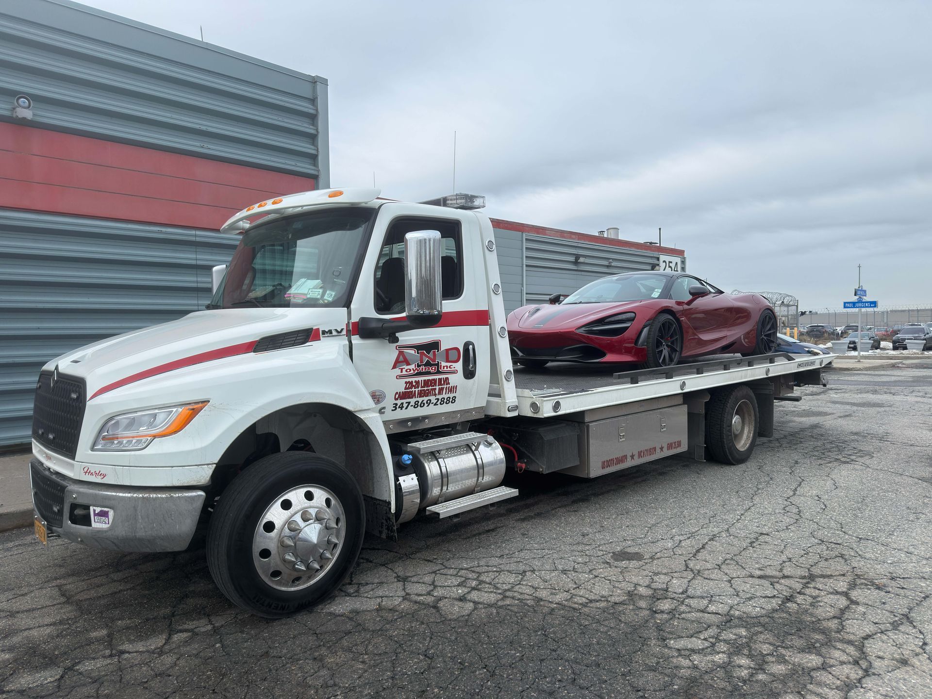 Tow truck carrying a red sports car in a gravel lot under cloudy skies