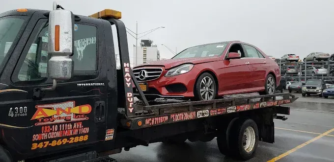 A red Mercedes sedan is on a flatbed tow truck. The tow truck is black, and the setting appears to be a junkyard.