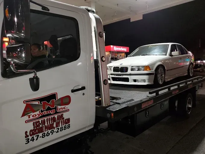 White BMW sedan being towed onto a flatbed tow truck at night, parked near a gas station.
