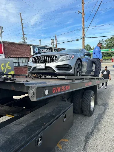 A gray car on a flatbed tow truck. Two men stand beside it on a sunny day.