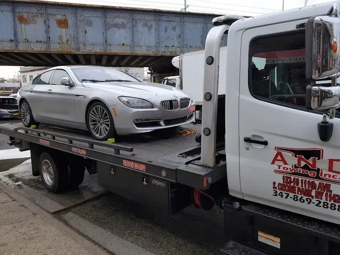Silver BMW car being towed on a flatbed tow truck beneath a bridge.