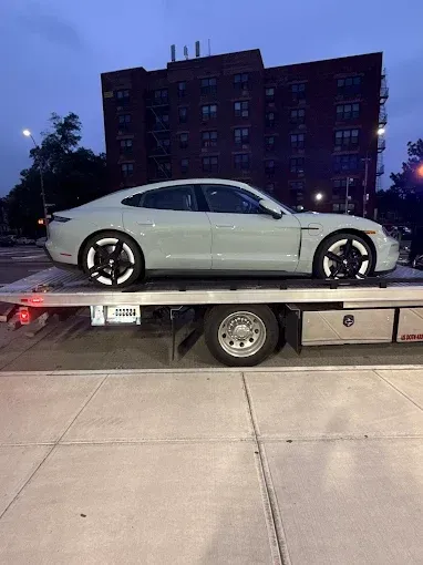 A light gray Porsche Taycan on a tow truck, parked on a city street at dusk.