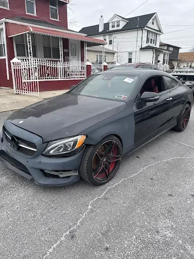Damaged black Mercedes-Benz coupe parked on a street; missing front bumper and fender, red rims.