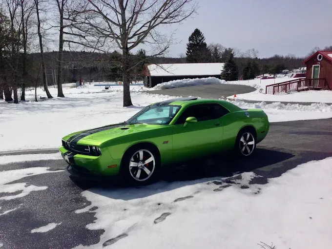 Green Dodge Challenger parked on a snowy driveway with bare trees and a red barn in the background