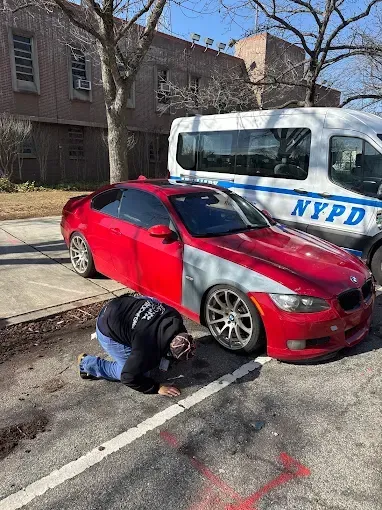 NYPD van by red sedan; person kneels beside the car in a parking lot.