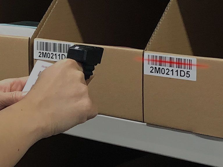 A person is using a barcode scanner (Hyco Ring Scanner) to scan a barcode on a cardboard box.