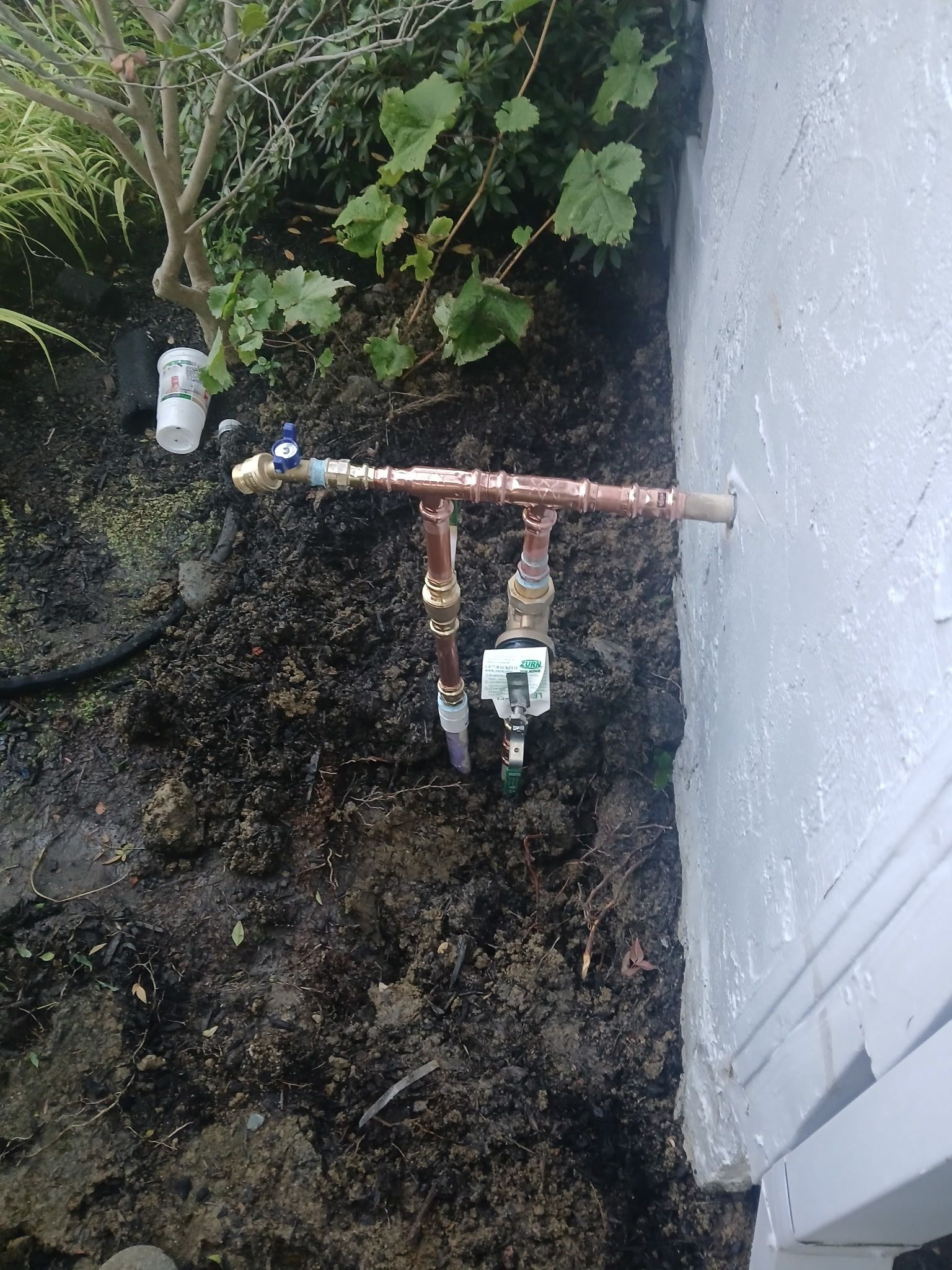 Rusty outdoor water pipes and meter beside a white wall, on muddy ground near plants