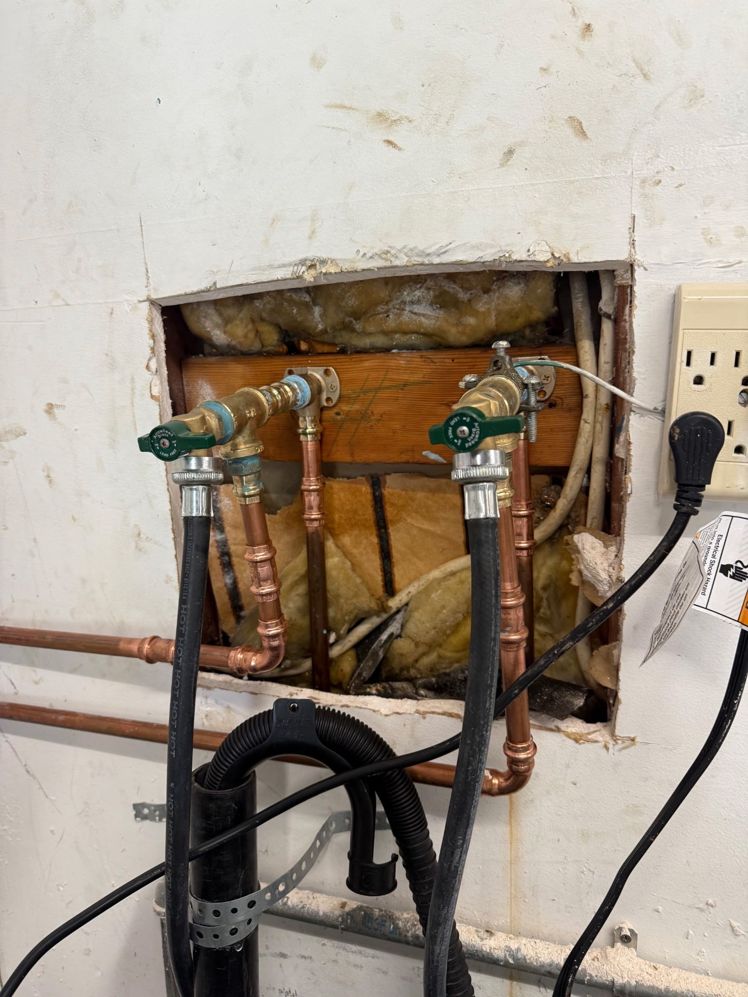 Exposed copper pipes and shutoff valves in a wall opening beside an electrical outlet with a black cable
