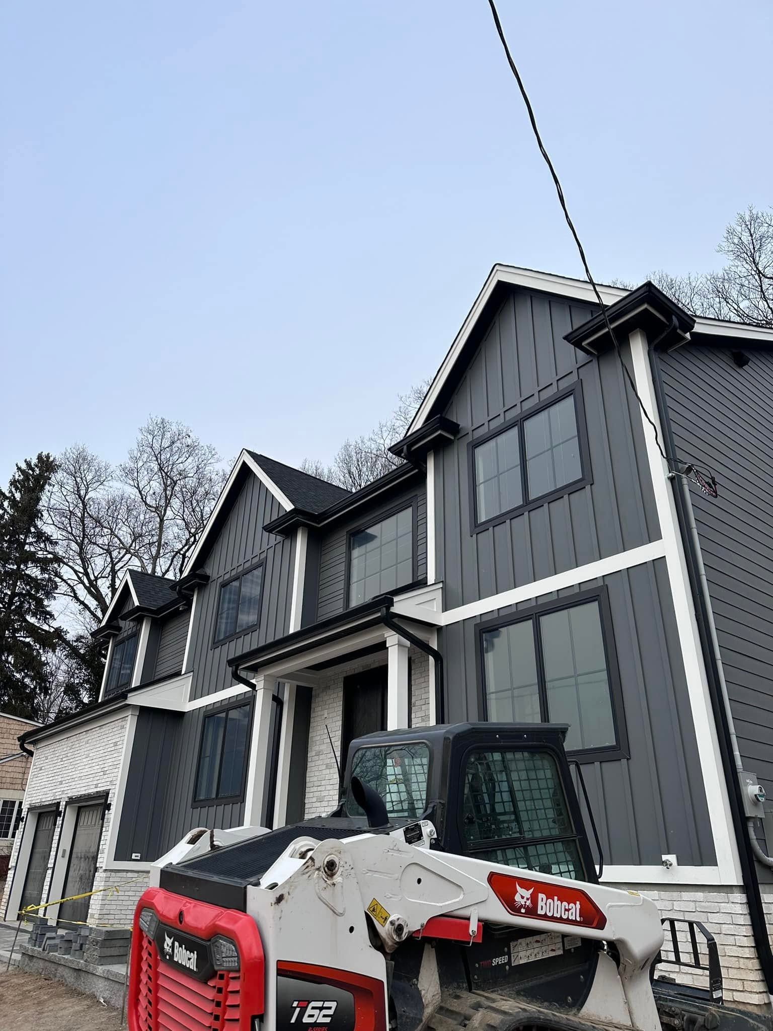 Snow-covered house with gray siding and a red-white Bobcat skid steer in the driveway