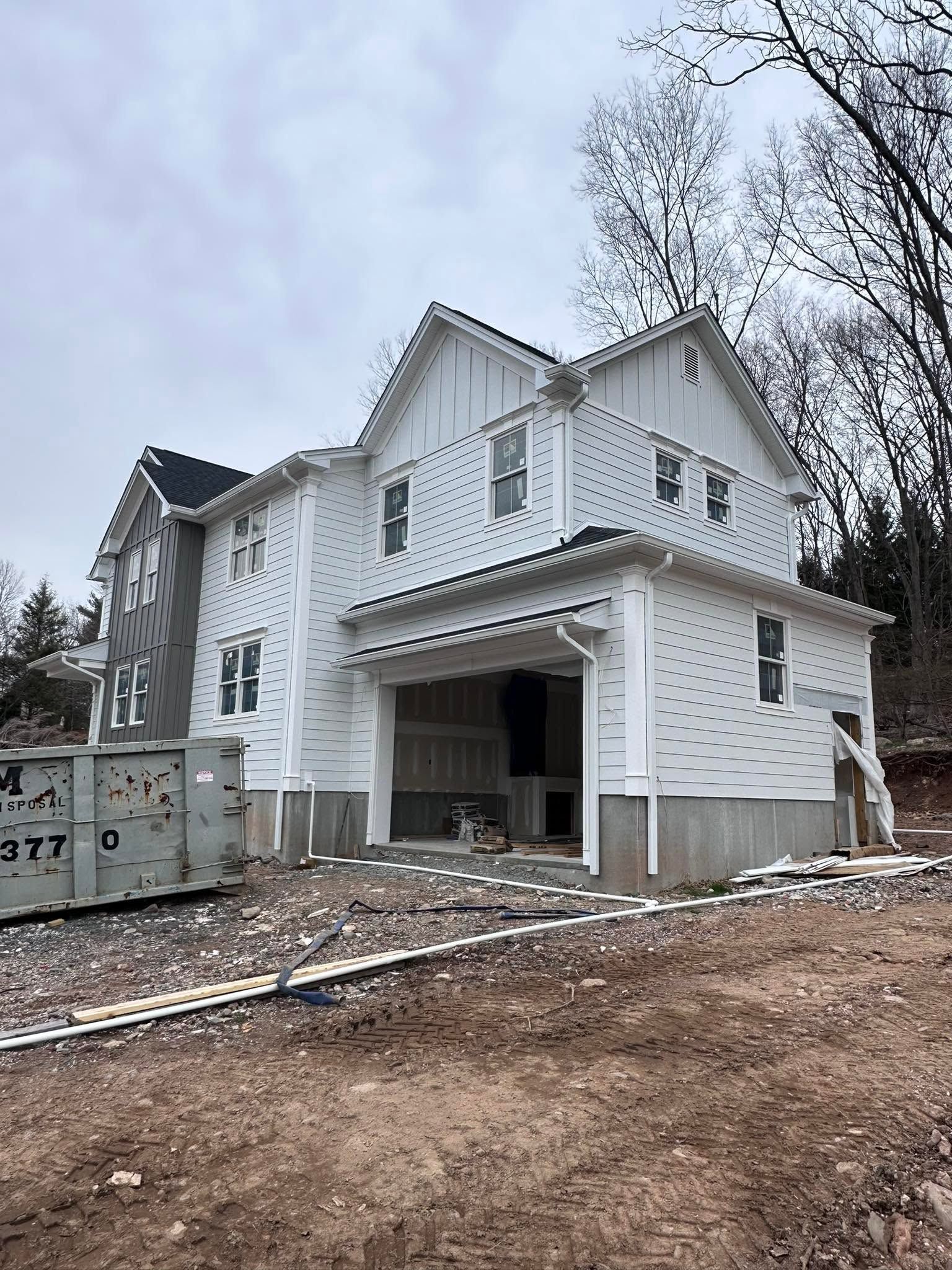 White two-story house under construction with open garage and dirt yard under cloudy sky