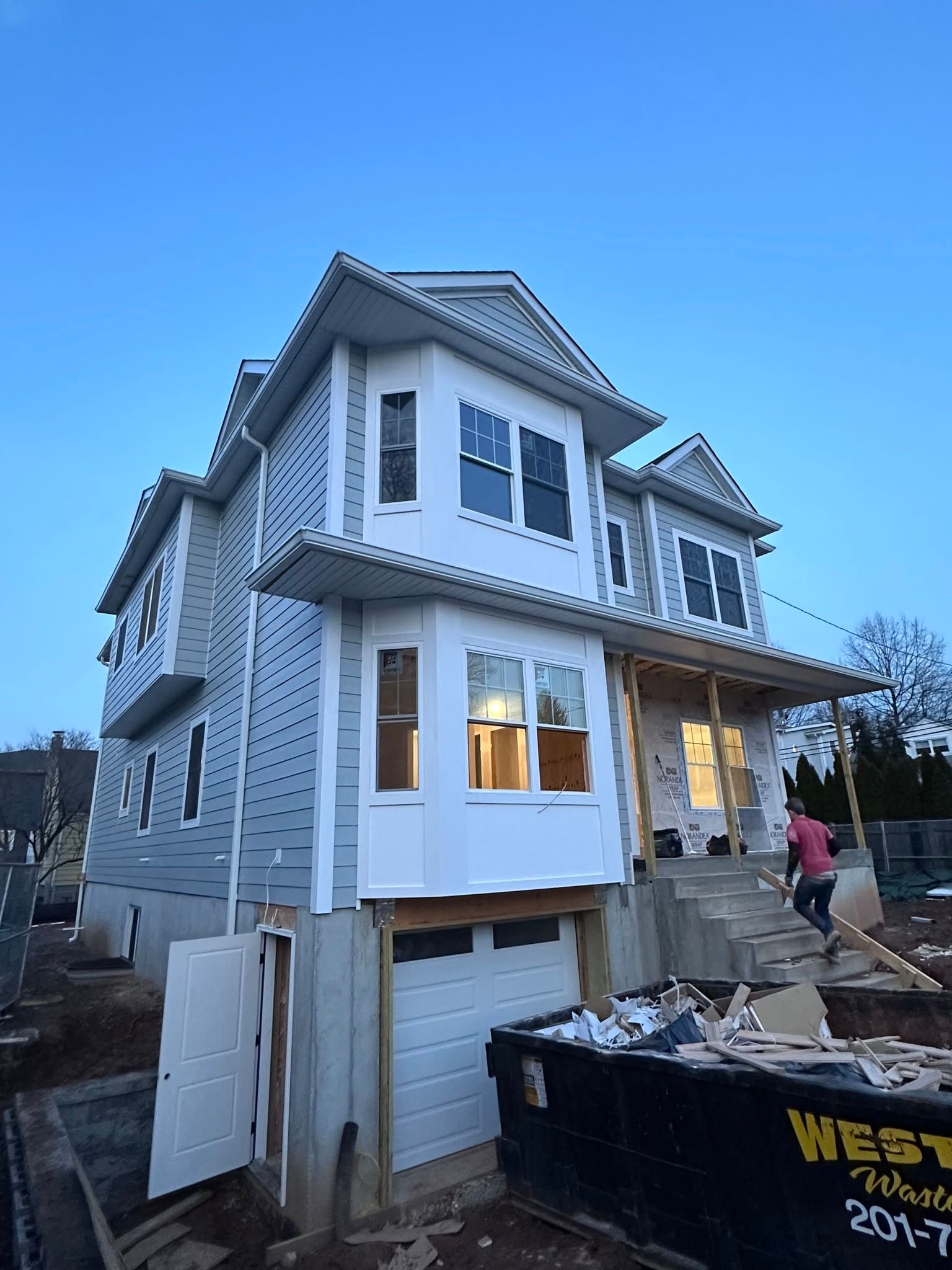 Two-story house under construction with white siding, garage below, and workers near a dumpster at dusk