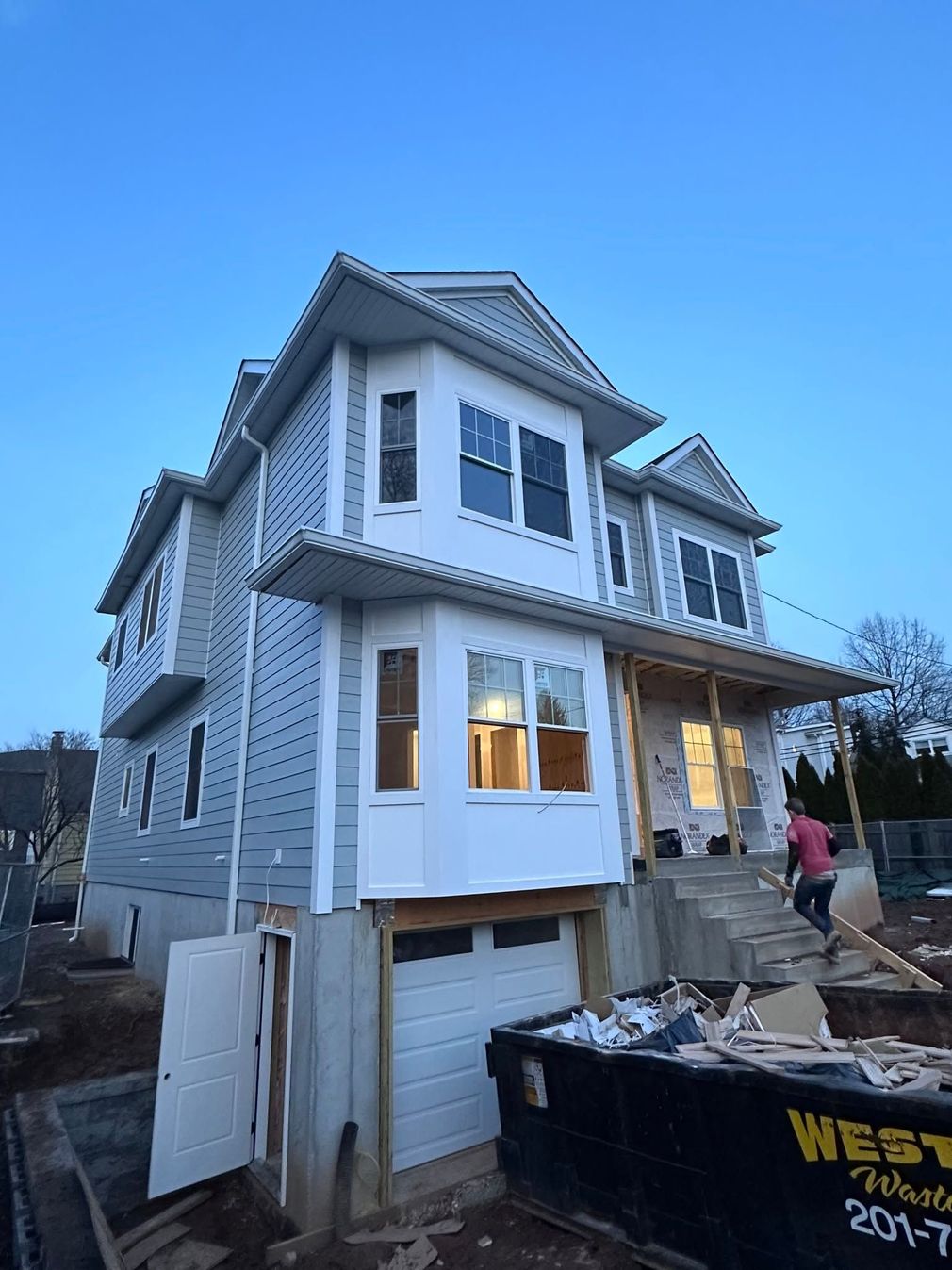Two-story house under construction with white siding, open garage, debris dumpster, and worker outside at dusk