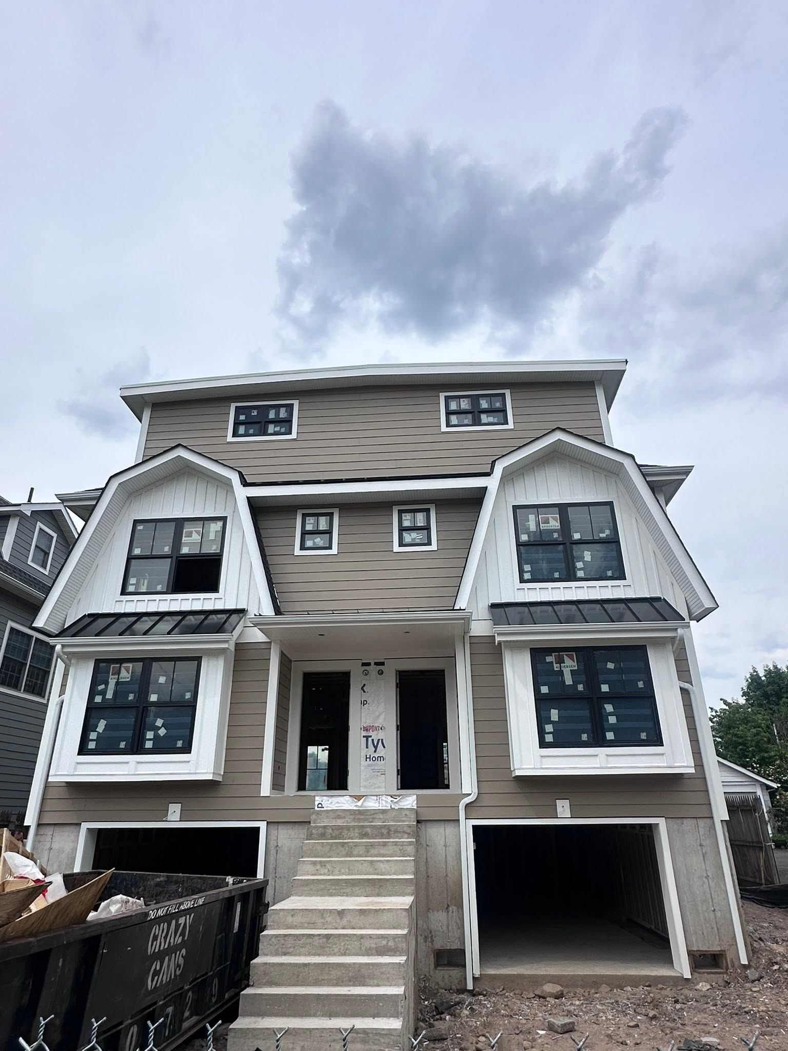 Front view of a multi-story house under construction with unfinished siding, stairs, and open garage bays