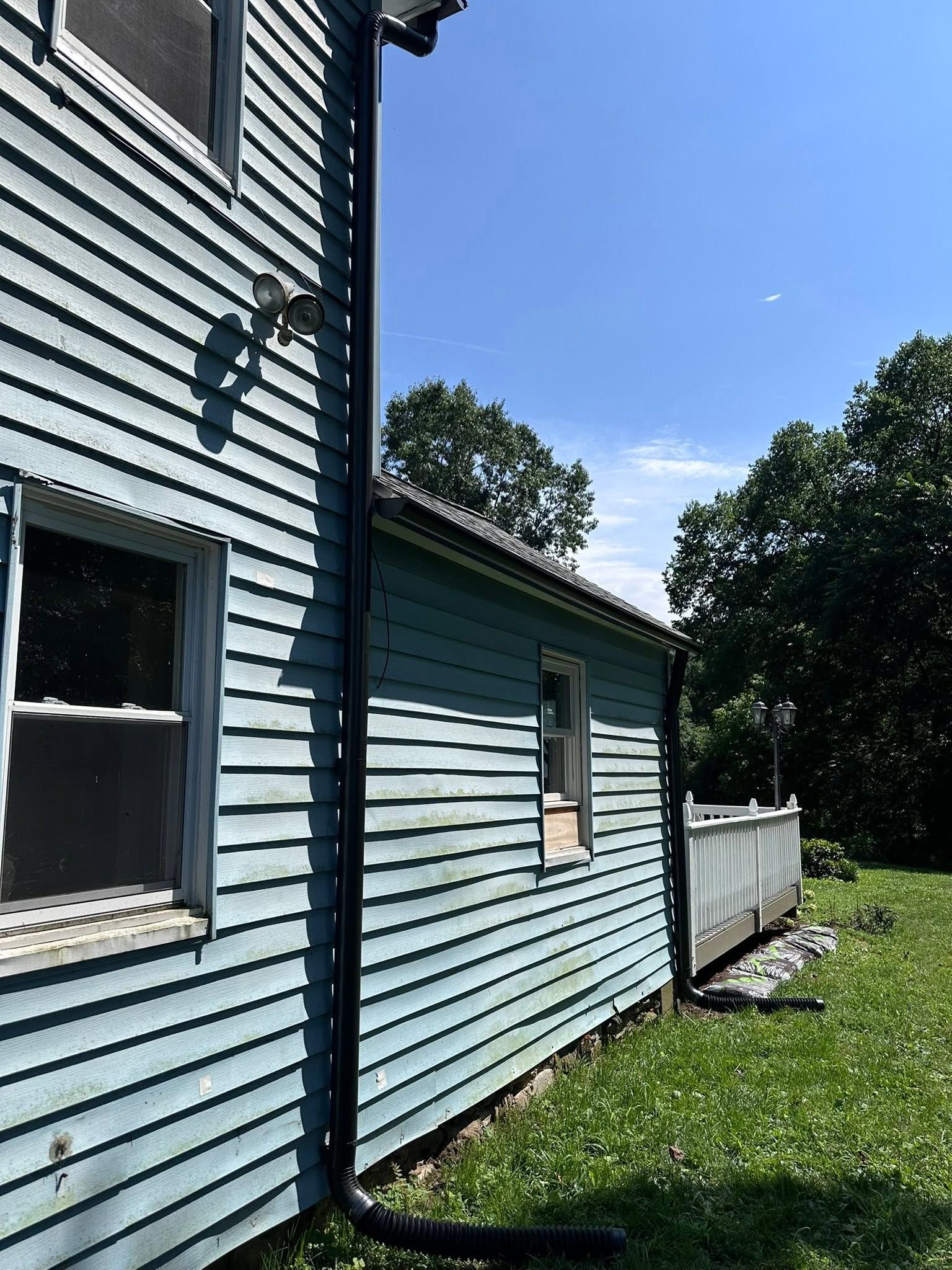 Blue house siding with windows, a downspout, and a white fence beside a grassy yard under a clear sky