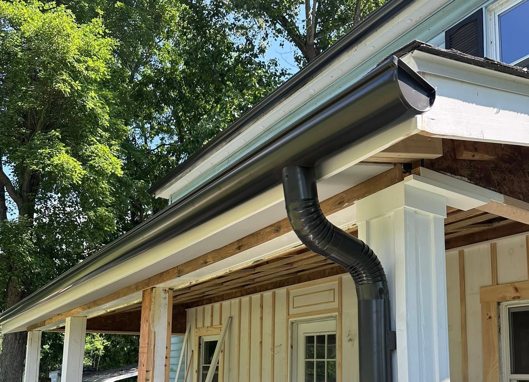 House eaves with black rain gutter and downspout above a porch, framed by trees.