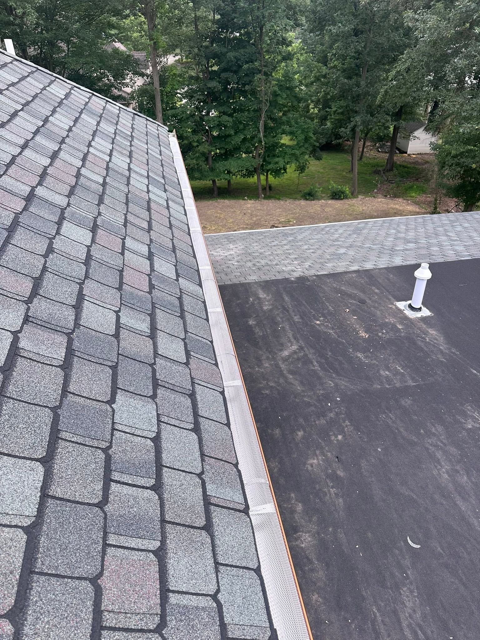 Roof shingles and a flat roof section beside trees, with a white vent pipe visible.
