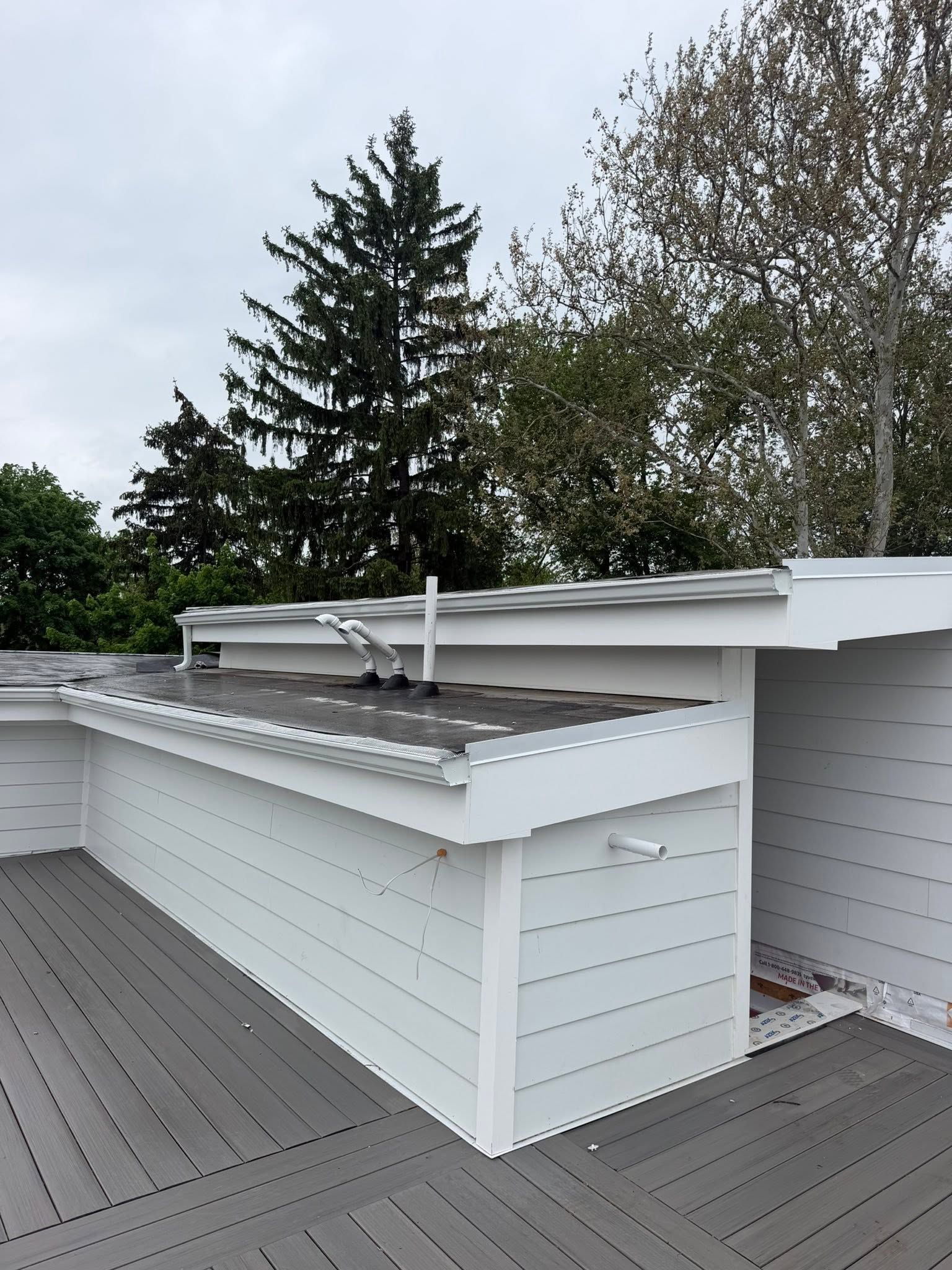 Rooftop deck with white railings and gray flooring beside a house, with trees in the background