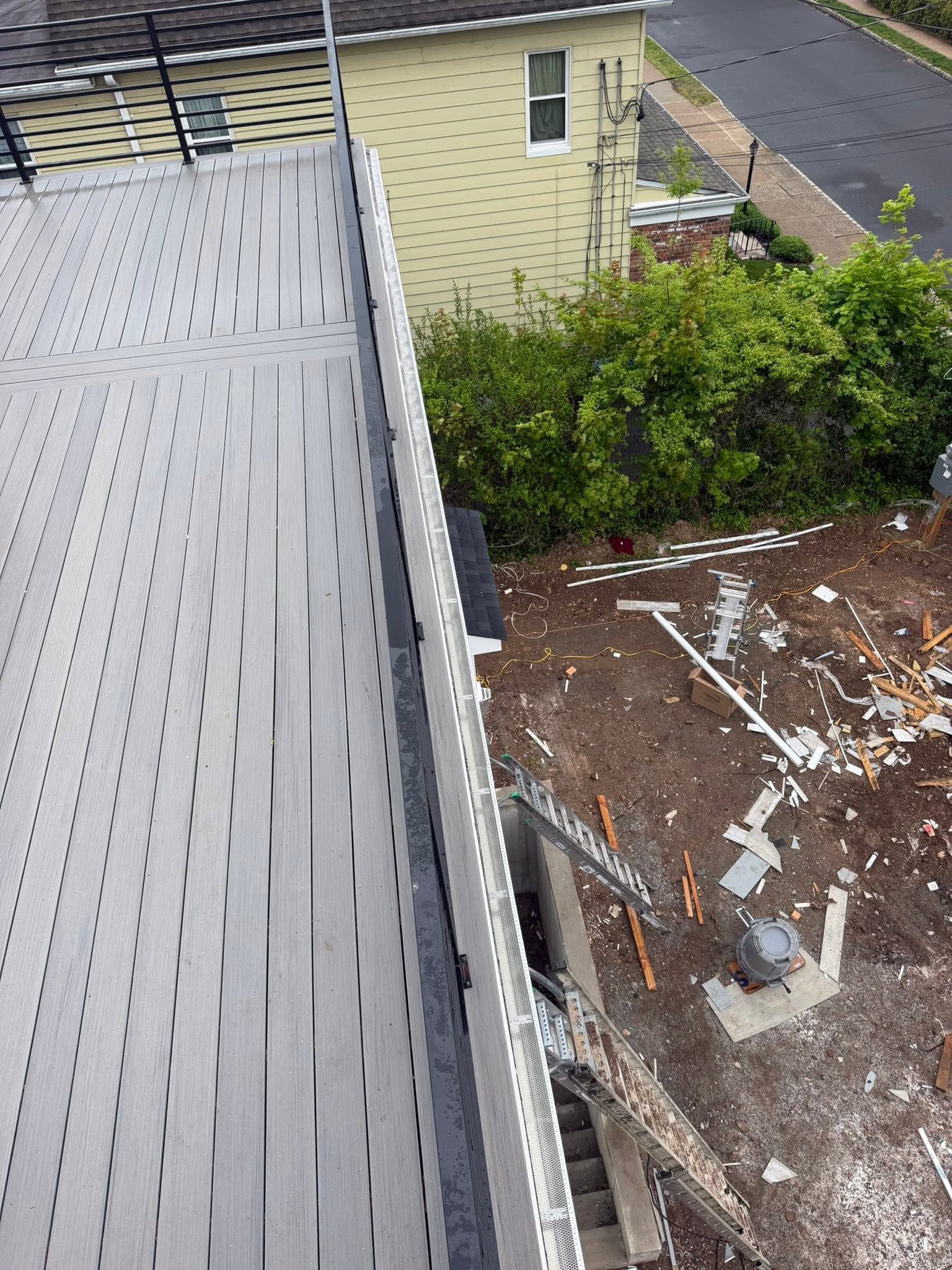 View from a rooftop: gray shingles, gutter edge, and a yard below with debris and dense green bushes