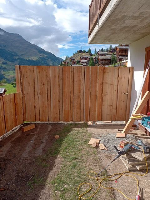 Une clôture en bois est en cours de construction devant un bâtiment avec des montagnes en arrière-plan.